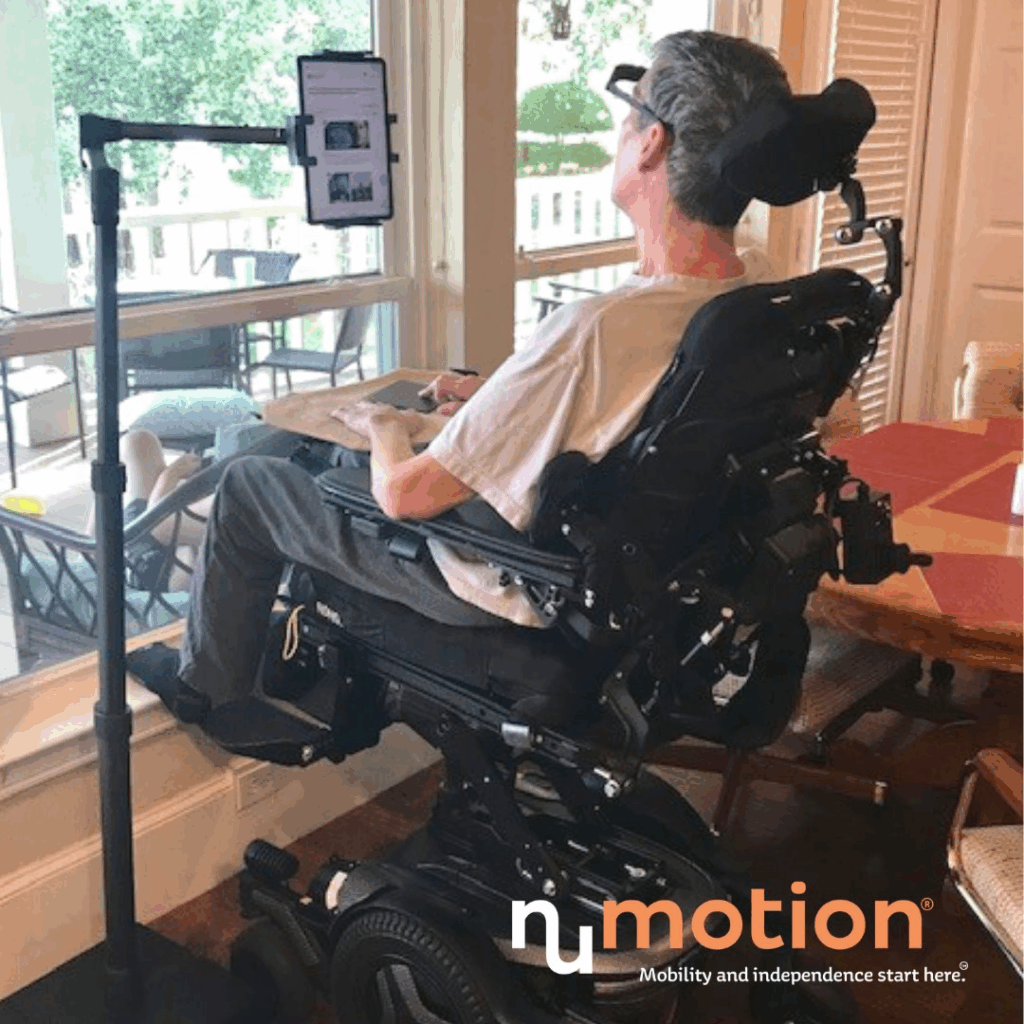 A man in a power wheelchair looks at a tablet mounted on a stand in front of him indoors by a large window, with the NuMotion logo visible in the bottom right corner.