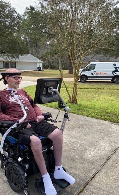 Kevins Story: A man in a motorized wheelchair with assistive technology sits outside on a driveway, wearing a maroon shirt, black shorts, and white socks. A NuMotion van is parked on the grass in the background.