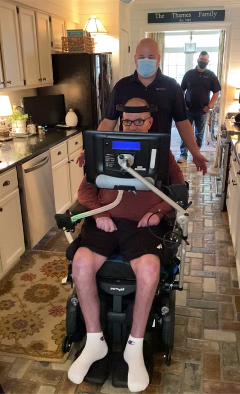 Kevin’s Story: A man using a powered wheelchair with assistive technology is in a kitchen with two men wearing face masks. The kitchen features white cabinets, brick flooring, and warm lighting.