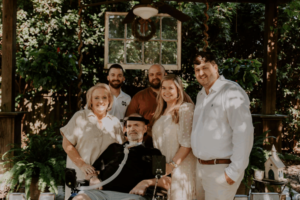 A group of six people, including an older man, pose and smile together outdoors in a shaded garden area with greenery and wooden structures in the background, sharing warmth as part of Kevins Story.