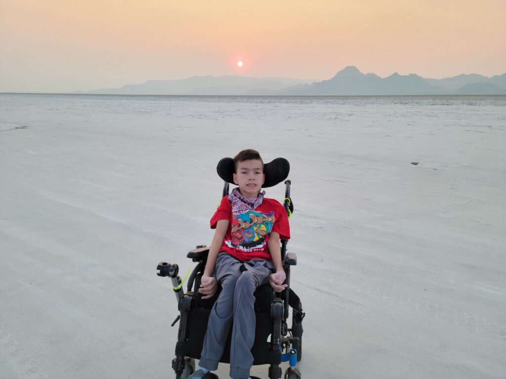 A boy, central to Xaviers Story, sits in a wheelchair on a vast, flat, sandy landscape at sunset. He wears a red T-shirt and gray pants, with distant mountains and an orange sun glowing in the hazy sky behind him.