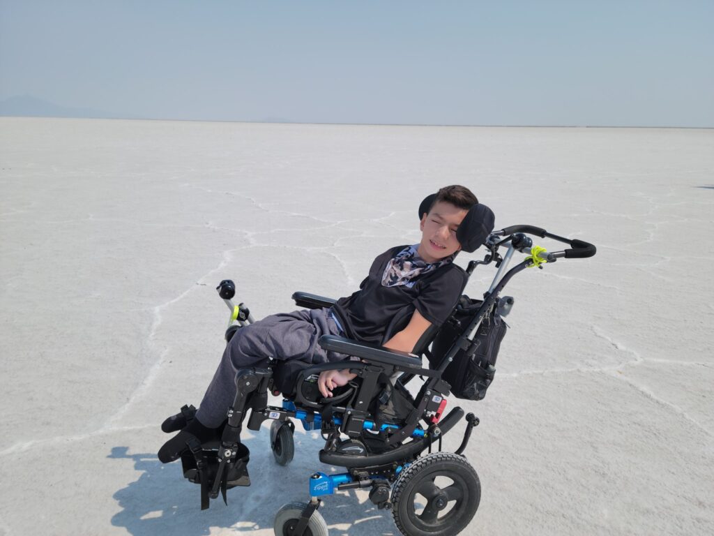 A boy in a powered wheelchair sits on a vast, flat, white salt flat under a clear sky, with distant mountains barely visible—capturing a moment from Xaviers Story.