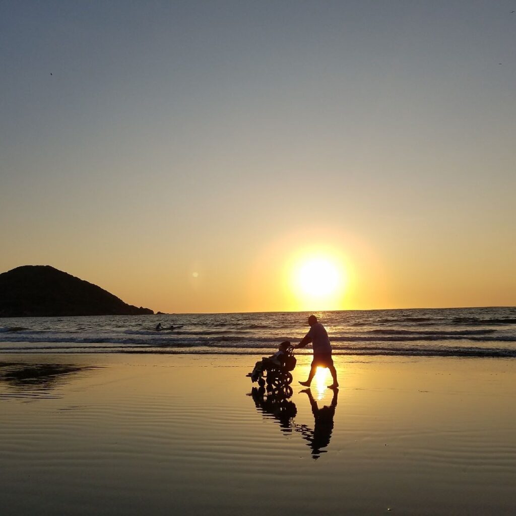 Xaviers Story unfolds as a person pushes a stroller along the beach at sunset, the low sun casting their silhouettes onto the shimmering wet sand, with a small hill in the background.