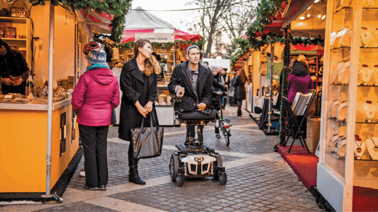 People browse stalls at an outdoor market decorated with garlands during Black Friday shopping. A man in a mobility scooter and a woman walk together, while other shoppers look at jewelry and gifts on display.