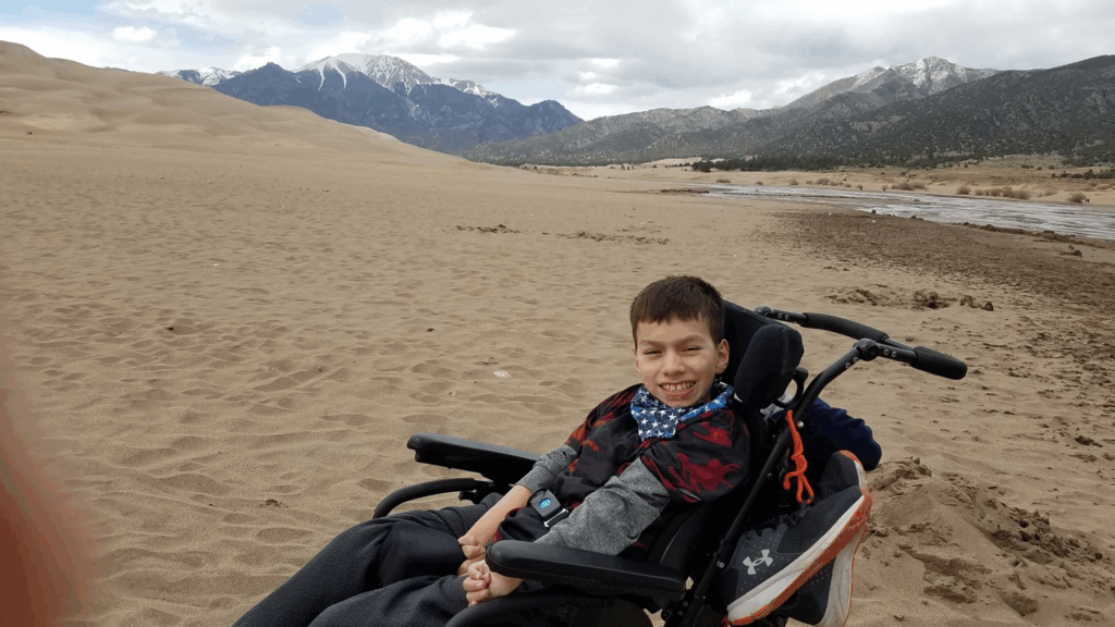 A smiling boy sits in a wheelchair on sandy terrain with mountains in the background and a river nearby under a cloudy sky, capturing a moment from Xaviers Story.