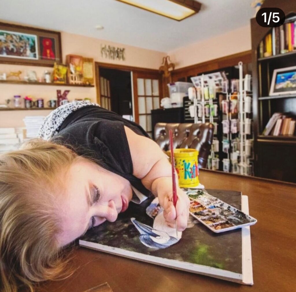 Katherines Story: A woman with a limb difference uses her mouth to paint on a canvas at a desk in a room filled with art supplies, books, and shelves.