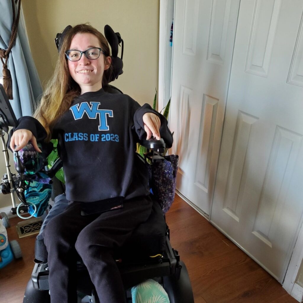Olivias Story: A young woman smiles at the camera, wearing glasses and a black WT Class of 2023 sweatshirt. She is seated indoors, near closed white closet doors and a wooden floor.