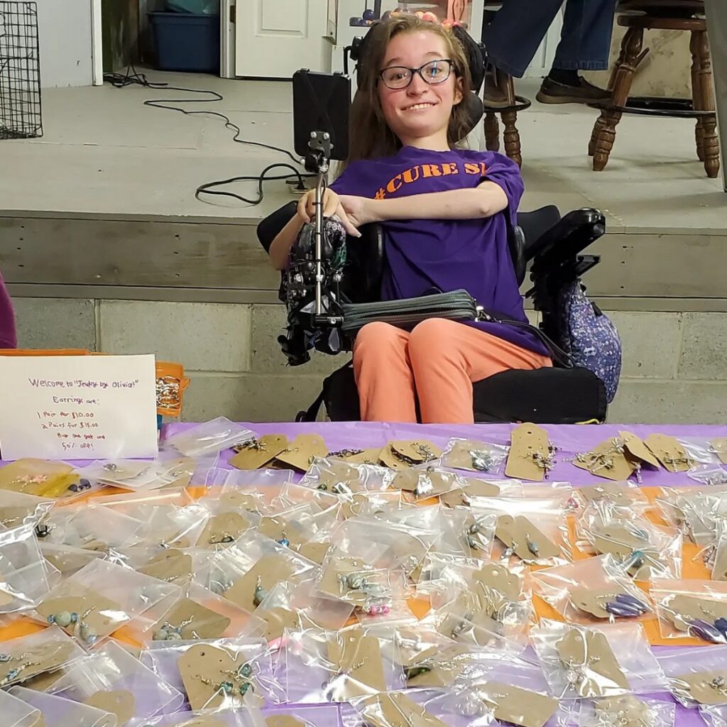 A smiling young woman sits behind a table covered with handmade jewelry in plastic bags. She wears glasses, a purple shirt, and orange pants, with a sign describing the items on display—an inspiring scene from Olivias Story.