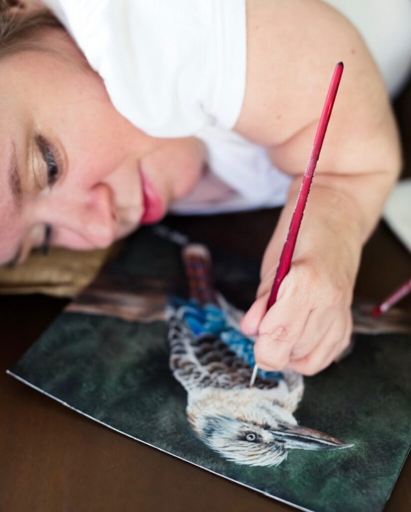 A woman lies on her side, carefully painting a detailed bird, possibly a kookaburra, on dark paper with a red-handled brush. This moment captures the artistry and dedication found in Katherines Story.