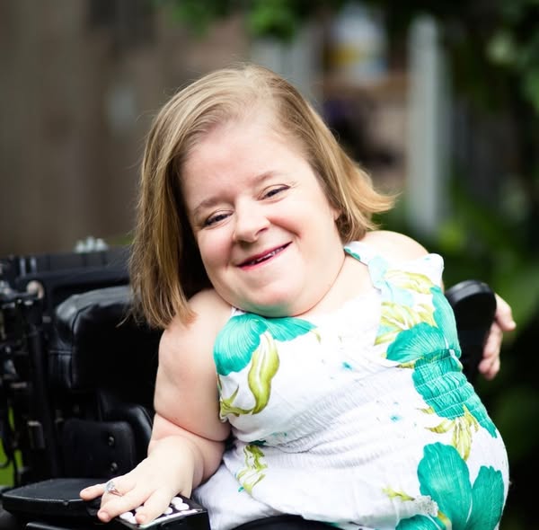Katherines Story: A woman with short blonde hair in a white floral dress sits in a wheelchair outdoors, smiling warmly at the camera with greenery blurred in the background.