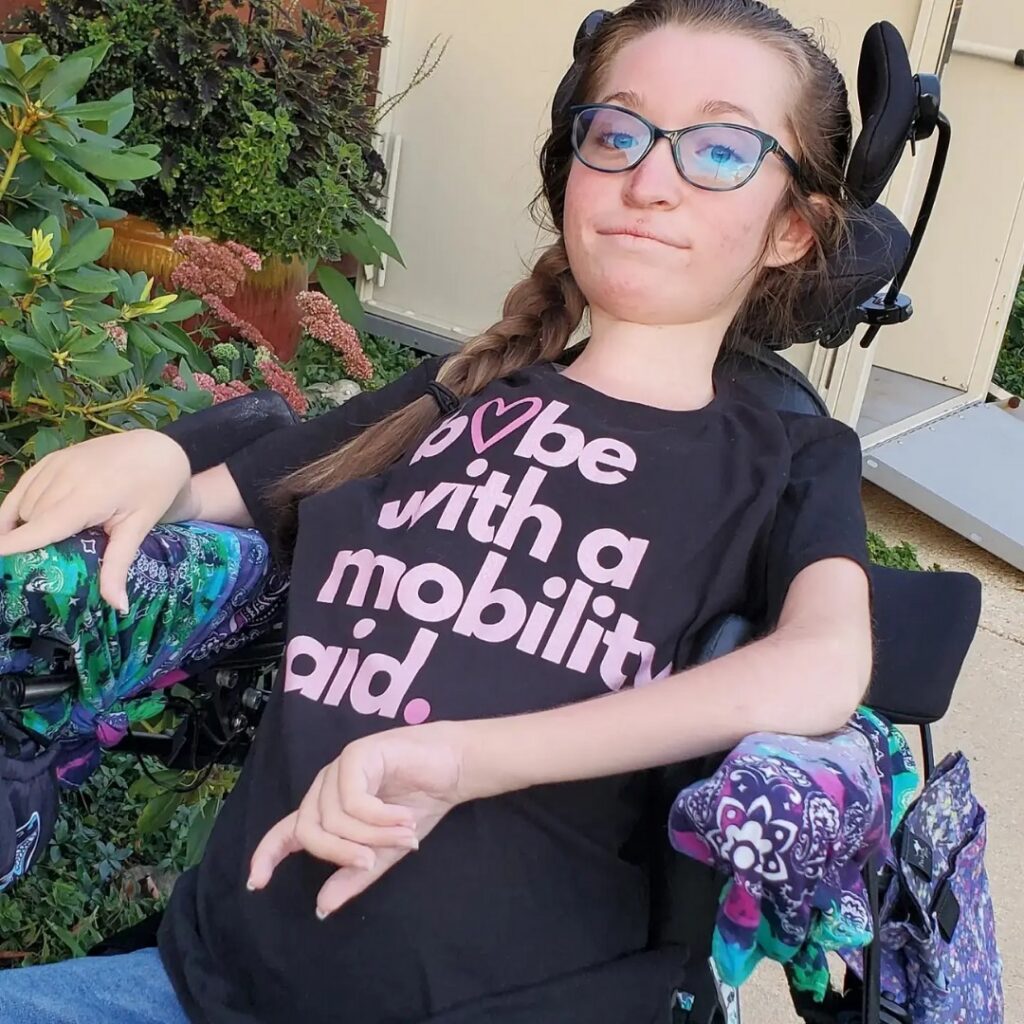 A young woman with glasses sits in a wheelchair, smiling softly. She wears a black t-shirt that says be with a mobility aid in bold letters, and patterned gloves. Green plants and a building frame Olivias Story in the background.