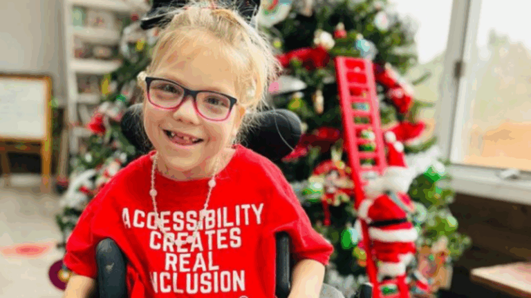 A smiling young girl with glasses and a wheelchair wears a red shirt that reads, ACCESSIBILITY CREATES REAL INCLUSION. She sits in front of a decorated Christmas tree, embodying the spirit of holiday season tips for creating real inclusion.
