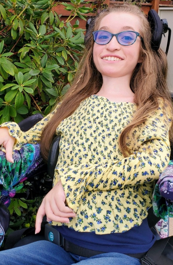 A young woman with long brown hair, wearing blue glasses and a yellow floral top, smiles while sitting in a wheelchair outdoors in front of green leafy bushes—capturing a joyful moment from Olivias Story.
