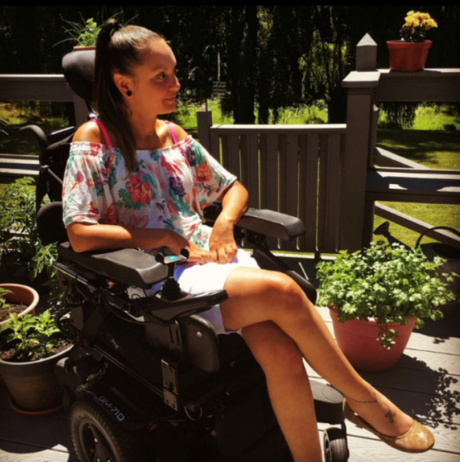 Morgans Story: A woman in a power wheelchair sits on a sunlit patio surrounded by potted plants. Wearing a floral top, white shorts, and tan flats, she smiles to the side with trees and greenery in the background.