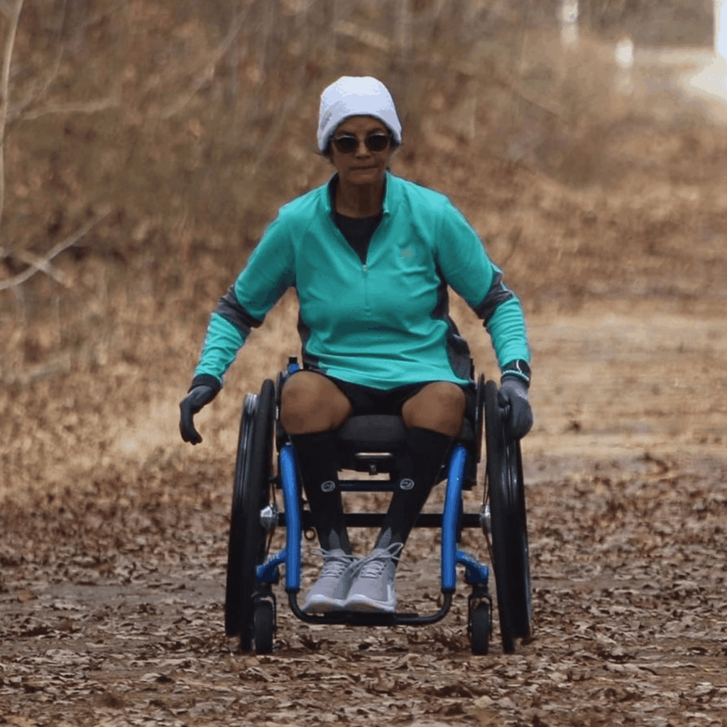 A woman in a wheelchair rolls along a wooded trail covered with dry leaves. She wears athletic gear, including a teal zip-up, black shorts, gloves, and a white beanie, with a focused expression and sunglasses.