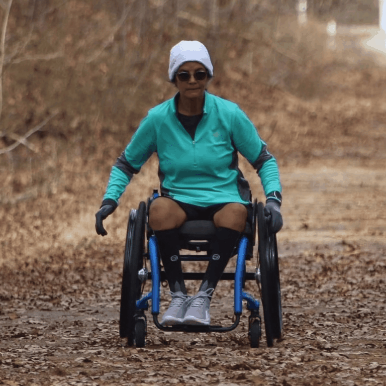 A woman in a wheelchair rolls along a wooded trail covered with dry leaves. She wears athletic gear, including a teal zip-up, black shorts, gloves, and a white beanie, with a focused expression and sunglasses.