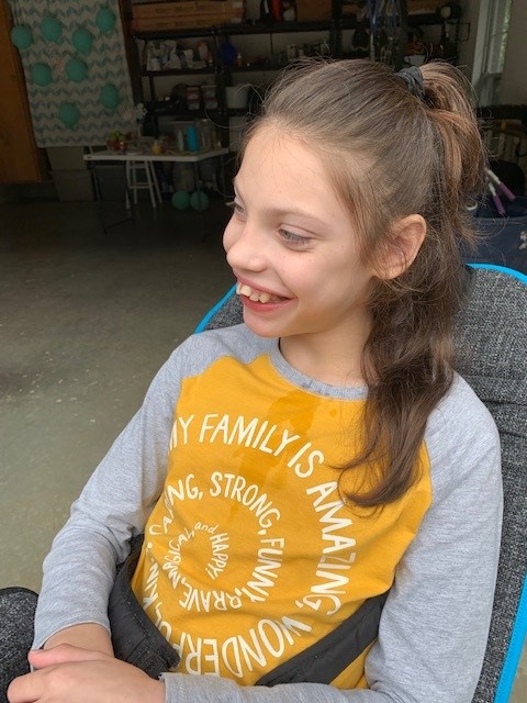 A smiling young girl with long brown hair in a ponytail sits in a chair, wearing a yellow and gray shirt that says MY FAMILY IS AMAZING, STRONG, FUN, AND WONDERFUL. She looks to the side in a garage setting, sharing Paulinas Story.