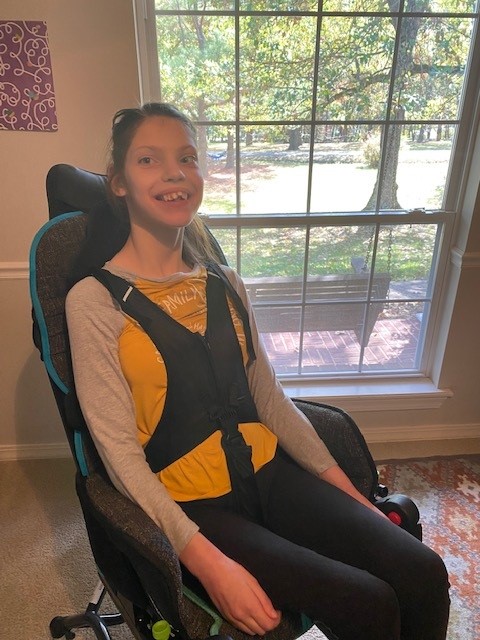 Paulinas Story: A young woman with long brown hair smiles while sitting in a wheelchair near a large window. She wears a yellow shirt with a gray cardigan and black harness, as sunlight and trees shine outside.