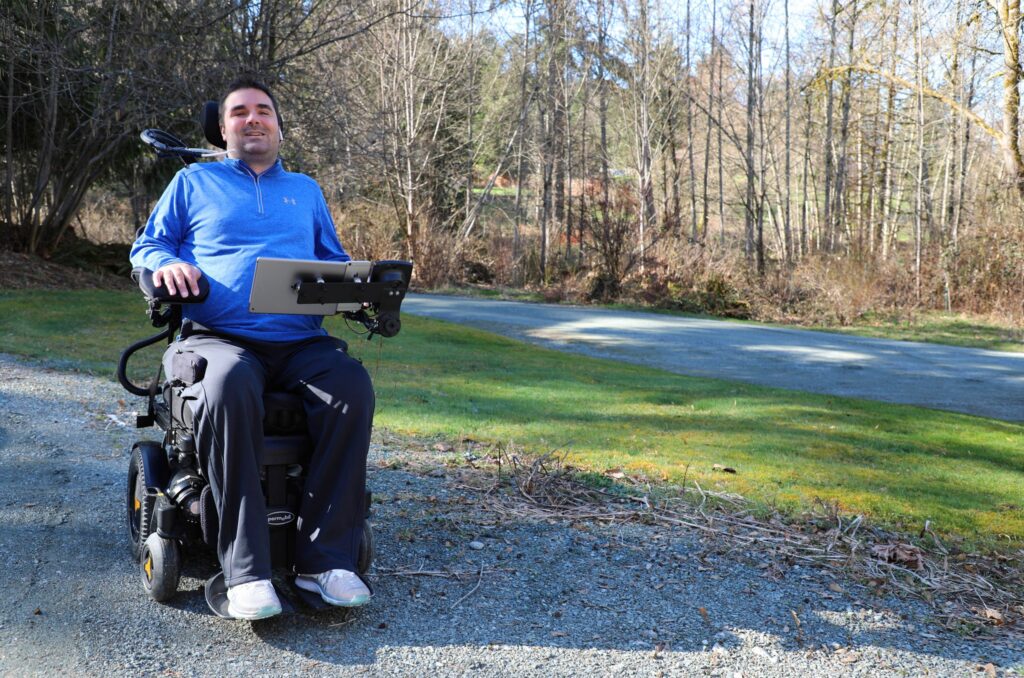 A man in a blue long-sleeve shirt sits in a motorized wheelchair with a tablet mounted in front of him, outdoors on a gravel path surrounded by grass and trees, sharing Tylers Story.