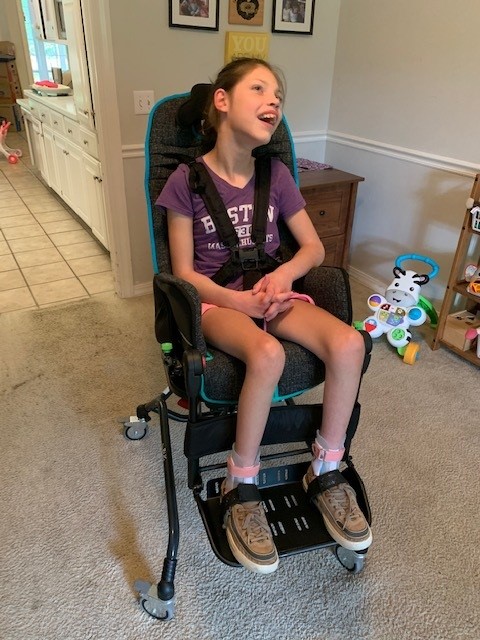 Paulinas Story: A young girl sits smiling in a supportive wheelchair with harnesses, wearing a purple T-shirt, shorts, and tan sneakers, in a bright room with toys and home furniture in the background.