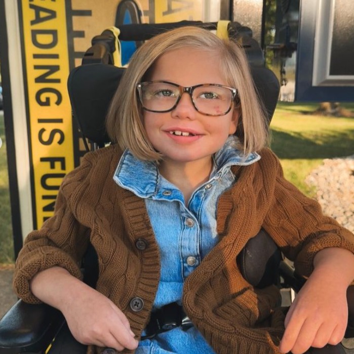 A young child with short blond hair and glasses sits in a wheelchair, smiling. Wearing a brown sweater over a denim shirt, they appear joyful. A yellow sign in the background reads Reading Is Fun, highlighting Evelyns Story.