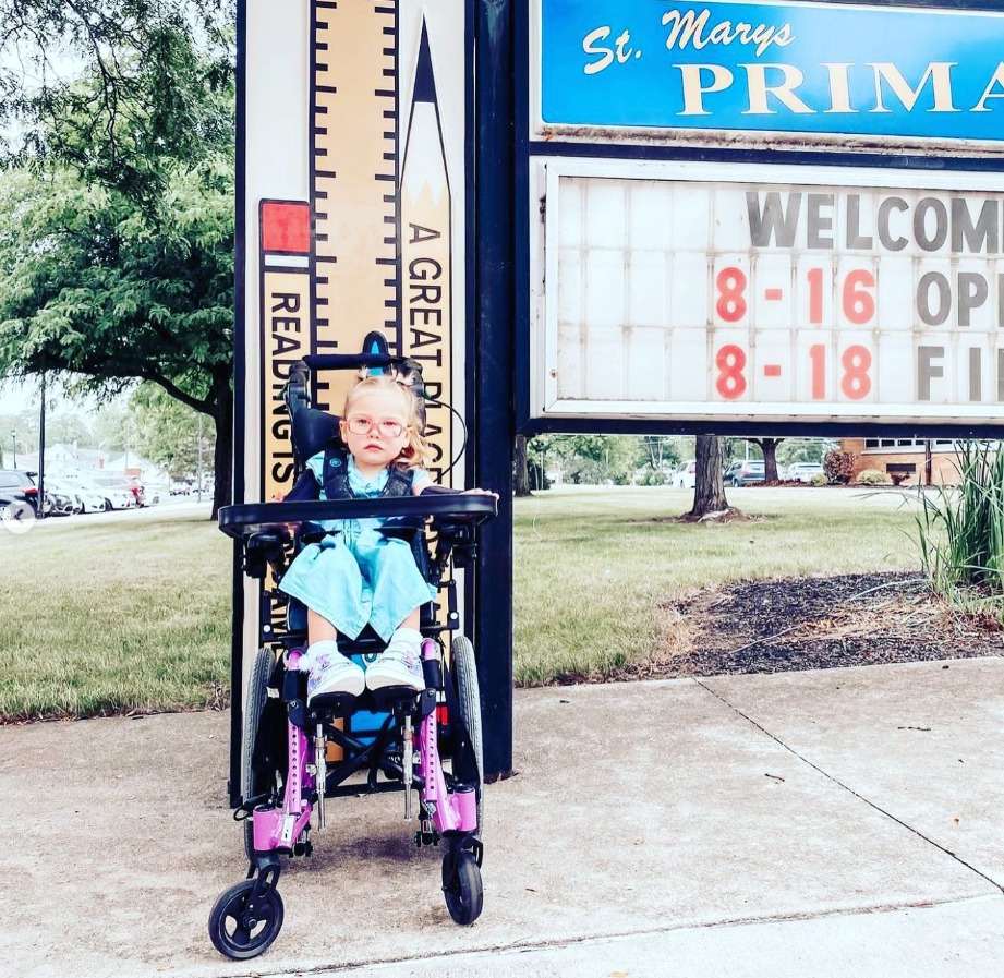 A young girl, part of Evelyns Story, sits in a wheelchair on a sidewalk before a school sign that reads WELCOME 8-16 OPEN 8-18 FIN. She wears glasses, a blue dress, and has a bow in her hair with trees and grass behind her.