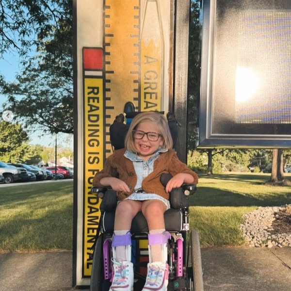 A young girl, featured in Evelyns Story, smiles as she sits in her wheelchair before a large decorative ruler and a Reading is Fundamental sign. She wears glasses, braces on her legs, and light clothes. Trees and parked cars are visible behind her.