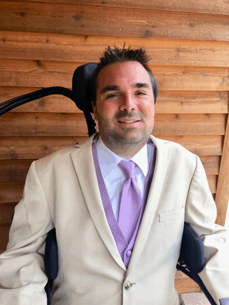 A man in a light tan suit, lavender tie, and purple vest smiles while sitting in a wheelchair in front of a wooden wall—part of Tylers Story.
