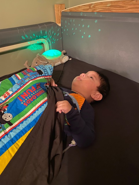 A young child lies in bed under a colorful blanket with train designs, looking up at glowing stars projected on the wall by a green nightlight. A stuffed animal rests nearby, part of Jakes Story.