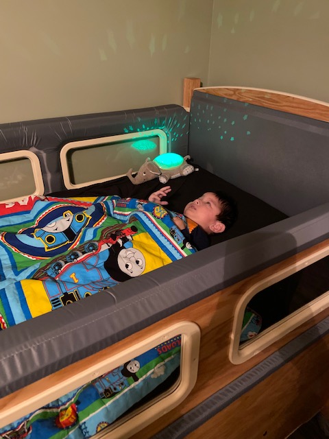 A young child sleeps in a wooden bed with gray bumpers, covered by a colorful Thomas the Tank Engine blanket. A green light projects stars on the wall, and a stuffed animal lays beside the child in Jakes Story.
