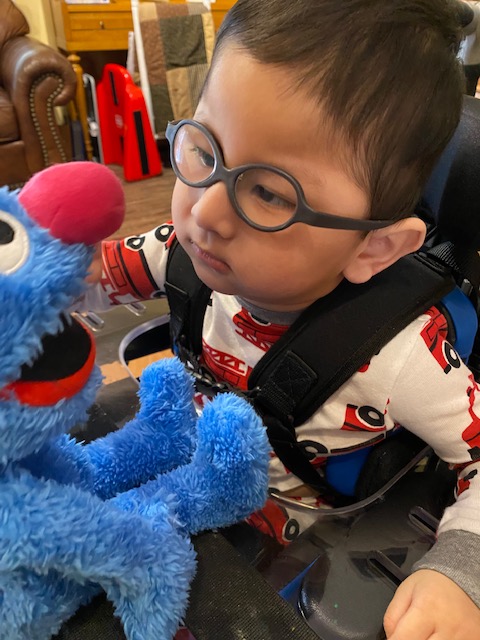 A young child wearing glasses and pajamas with red cars sits in a wheelchair, looking at a blue Grover puppet with a big pink nose and open mouth. This touching scene is part of Jakes Story, set indoors with furniture in the background.