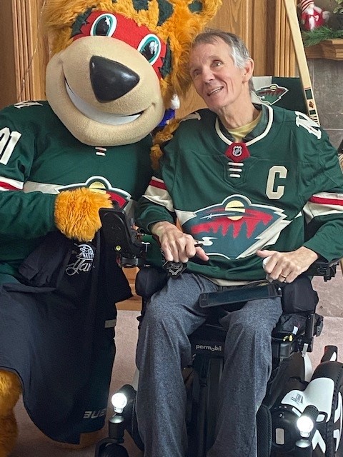 A man using a wheelchair, wearing a Minnesota Wild hockey jersey, smiles as he poses indoors next to the team mascot. Both appear happy and engaged in their matching jerseys.