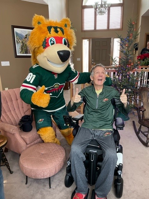 A person using a wheelchair smiles and gives two thumbs up next to a Minnesota Wild mascot indoors, near a Christmas tree and pink armchair.