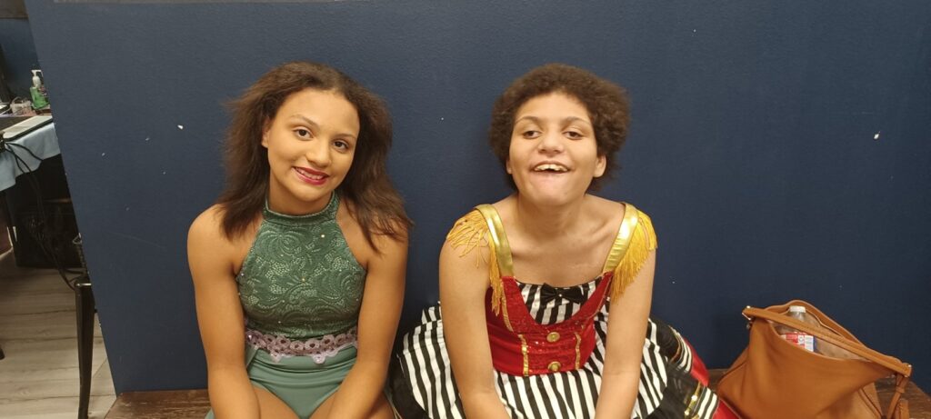 Two young women in colorful costumes sit side by side, smiling at the camera. Amayas Story unfolds as one wears a green lace outfit and the other a red, yellow, and black striped dress. A brown bag rests on the floor by a blue wall.