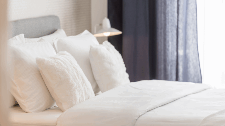 A neatly made bed with white linens and fluffy pillows, set next to a white lamp and dark curtains in a softly lit, modern accessible room—ideal for your next booking.