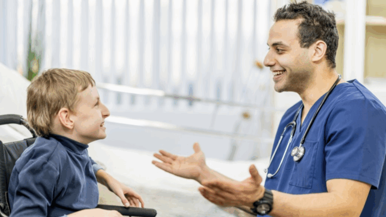 A doctor in blue scrubs with a stethoscope is smiling and talking to a young boy in a wheelchair. They are indoors, facing each other, both appearing happy and engaged in conversation about doctors visit preparation.