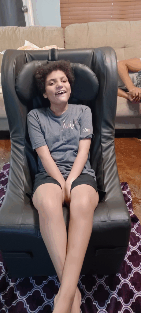 A smiling child with short curly hair sits in a black supportive chair, wearing a gray t-shirt and black shorts. Amayas Story unfolds as another person appears in the background, sitting on a beige couch, mostly out of frame.