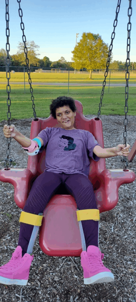 A smiling child with curly hair sits on an adaptive red swing at a park, wearing a purple t-shirt, maroon leggings, pink high-top shoes, and leg braces. Green grass and trees frame the background in Amayas Story.