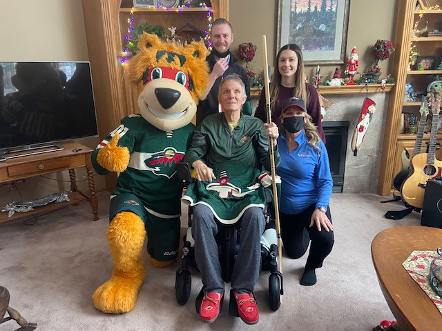 A group poses indoors, including a person using a wheelchair, two women, one man, and someone in a Minnesota Wild mascot costume. The room is decorated for Christmas with stockings, lights, and a tree.