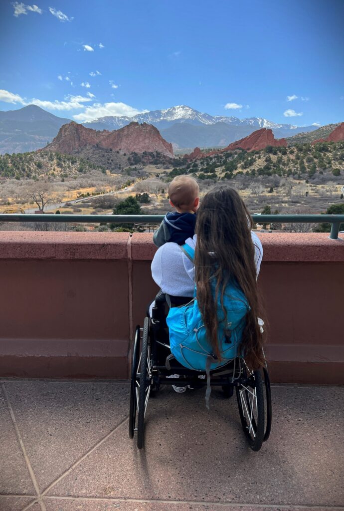 Elizabeth’s Story: A woman with long hair and a turquoise backpack holds a small child on her lap, both gazing at the scenic red rock formations and mountains under a blue sky.