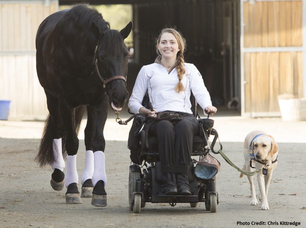 A smiling woman in a power wheelchair holds the leash of a yellow lab and leads a black horse. Featured in Stefanies Story, she is outside near horse stables as the horse and dog walk closely beside her.