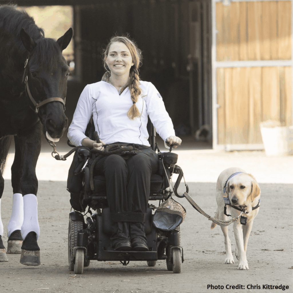 A smiling woman in a power wheelchair leads a black horse and a yellow service dog outside a stable. She wears a white riding jacket, and the animals walk calmly on either side of her.