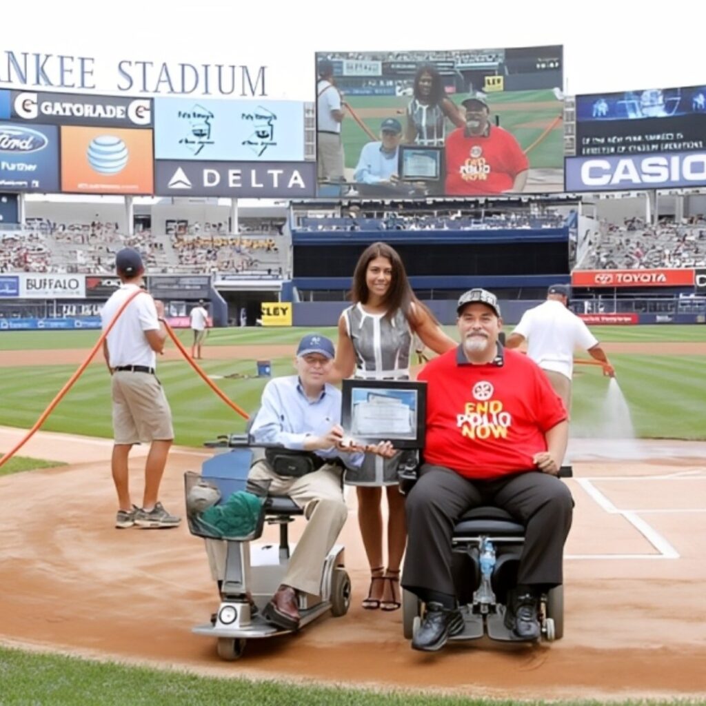Two men in motorized wheelchairs and a woman stand on the baseball field at Yankee Stadium, holding a certificate. A large screen behind them shows their image, celebrating John’s Story as workers prepare the field in the background.