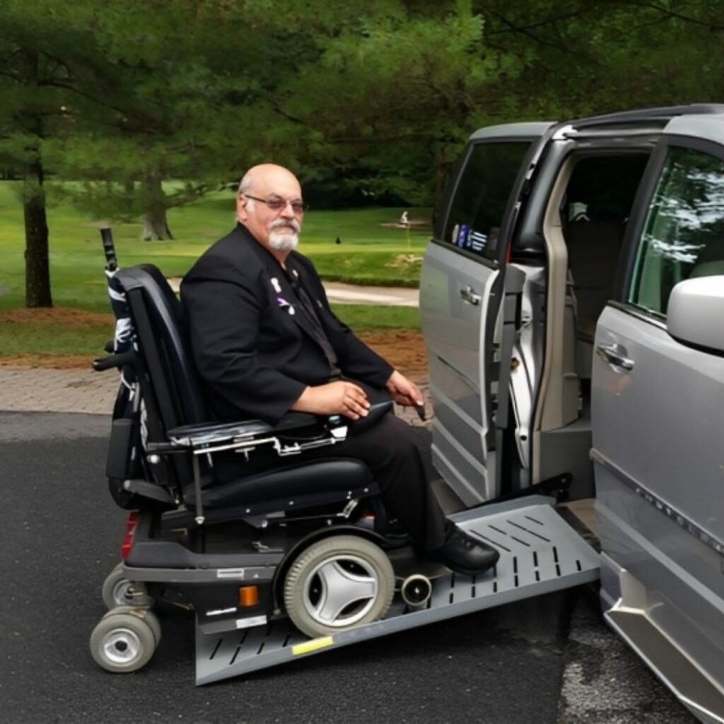 John’s Story: A man in a suit sits in a motorized wheelchair, using a ramp to enter a silver accessible van parked outdoors near trees and grass.