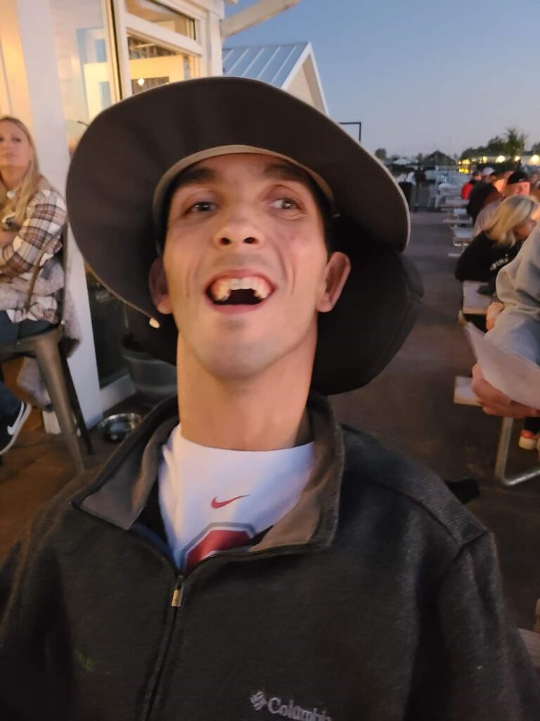 Rickys Story: A man wearing a wide-brimmed hat and a zip-up jacket smiles joyfully at an outdoor restaurant in the evening, with other people seated at tables in the background.