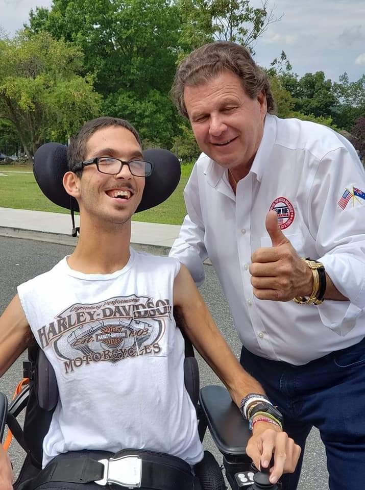 A smiling young man in a wheelchair wears a Harley-Davidson T-shirt while an older man in a white shirt stands beside him, giving a thumbs up. They are outside on a sunny day with trees in the background, celebrating Rickys Story.