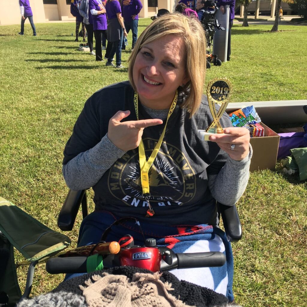 A smiling woman sits in a mobility scooter outdoors, holding and pointing to a small trophy labeled 2019. Moniques Story shines as she wears a gray shirt and yellow lanyard, with people and buildings in the background.
