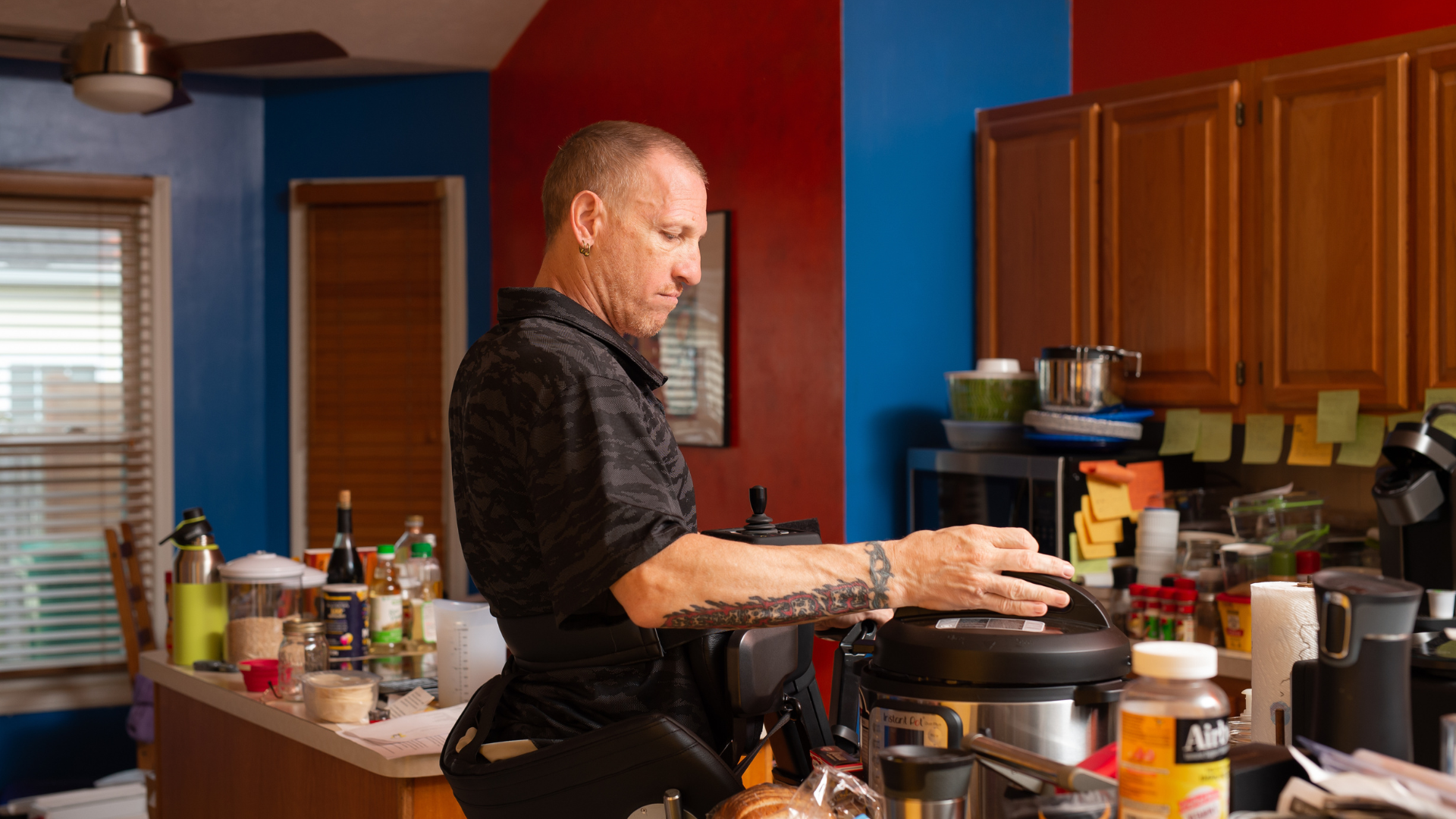 A man with a tattoo on his right arm prepares food in a cluttered kitchen, using a pressure cooker on the counter. Kitchen shelves and counters are filled with containers and ingredients, highlighting some accessibility tips for an accessible apartment.