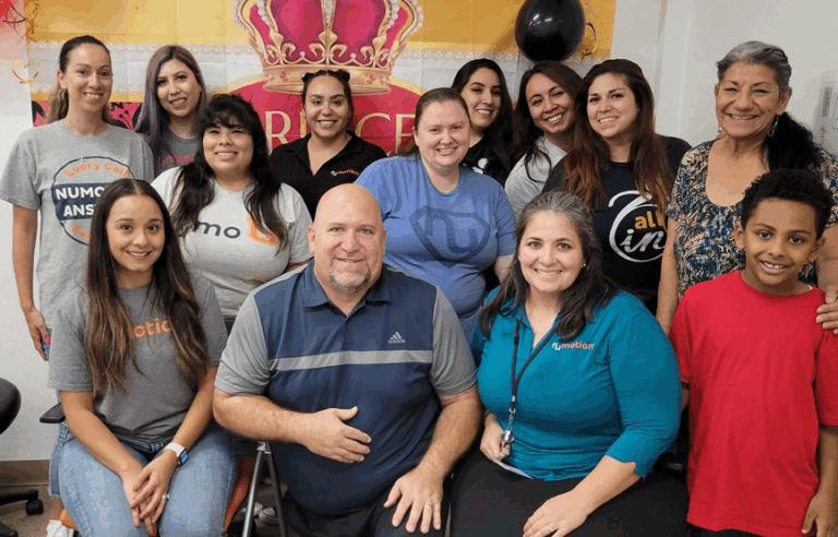 A group of thirteen adults and one child smiling and posing indoors in front of a Princess banner. The group includes men and women of various ages, most wearing casual clothing, gathered to celebrate Jeffs Story.