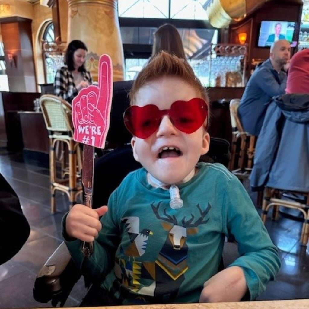 A smiling child wearing heart-shaped red sunglasses and a blue shirt with animal prints holds a foam finger that says We’re #1 while sitting in a restaurant. Noah’s Story shines as other people are seen in the background.